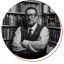 James Emanuel Shapiro Headshot, Waist up, man with white collered shirt and sweater vest standing in front of bookcase. Short black hair and rectangular glasses